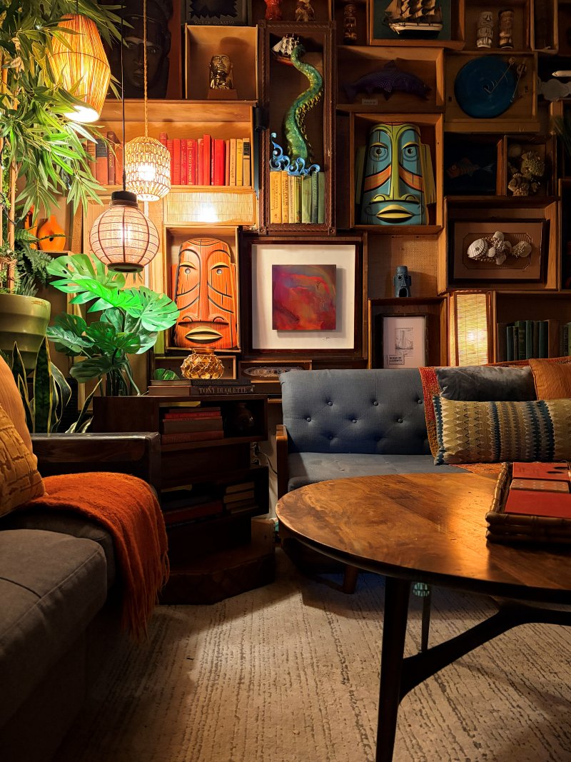 Client tiki lounge with a wall of carved masks, a blue tufted sofa, and rattan lanterns